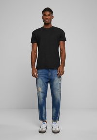 Camiseta de algodón negra combinada con jeans de mezclilla destrozados en azul claro y zapatillas blancas con detalles en negro y gris. El modelo posa de forma frontal.