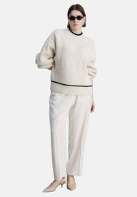 Elfenbeinfarbener, übergroßer, gerippter Pullover mit schwarzem Saum, kombiniert mit crema-farbenen, weiten Hosen und nudefarbenen, spitzen Heels; inklusive schwarzer Sonnenbrille.