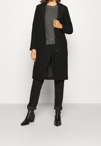 Cappotto di lana nero con finitura testurizzata, design monopetto e rever, indossato sopra un maglione grigio e pantaloni neri, abbinato a stivaletti.