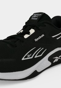 Αθλητικό παπούτσι Reebok για προπόνηση σε ασπρόμαυρο με κορδόνια, ορατή σήμανση στο πλάι, στη γλώσσα και στην σόλα, εμφανίζεται υπό γωνία σε ουδέτερο φόντο.