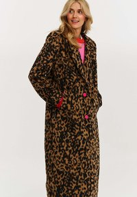Žena v dlhom kabáte s leopardím vzorom a ružovými gombíkmi, s rukami v vreckách, a s ružovým topom pod ním, pred jednoduchým pozadím.