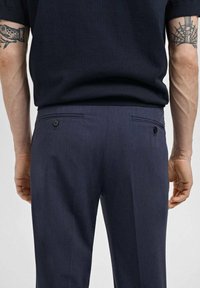 Pantalons pour hommes de couleur marine en tissu texturé avec deux poches arrière et des fermetures à bouton. Design ajusté, détails minimaux sur la taille.
