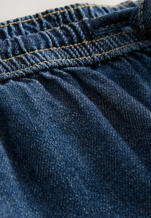 Denimstoff in Blautönen mit Gummizugbund. Auffällige sichtbare Nähte in kontrastierendem Goldton. Glatte Textur.