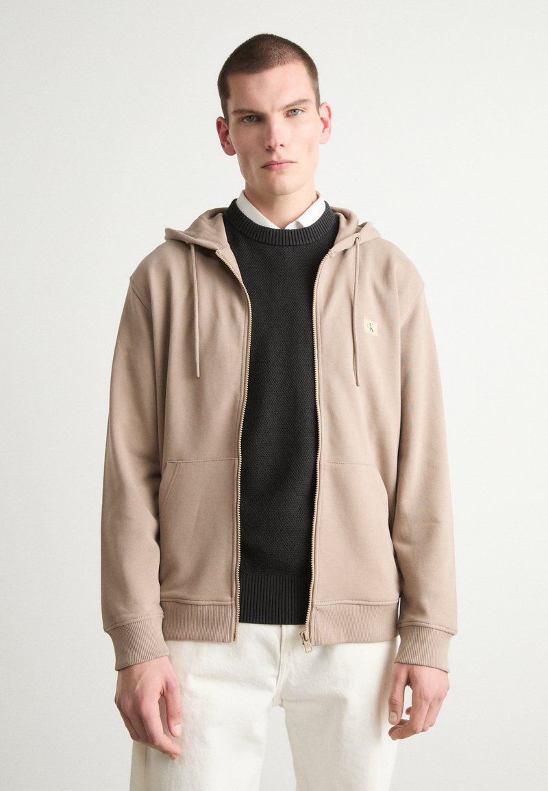 Felpa beige con zip, cappuccio regolabile, tasche frontali e un piccolo logo. Indossata sopra un maglione nero con texture e pantaloni bianchi. Vista in prospettiva accorciata.