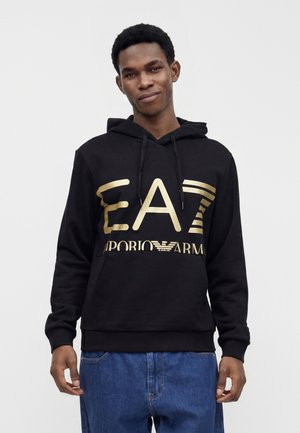 Jeune homme portant un sweat à capuche noir avec le logo doré "EA7 Emporio Armani" et un jean bleu, debout devant un fond clair uni.