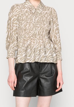 Femme portant un chemisier smocké beige et blanc à motifs avec des manches trois-quarts bouffantes et un short en cuir noir.