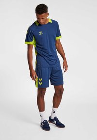 Navy blauwe sportoutfit met limoen groene accenten. Bevat een kort- mouwen top en shorts met chevronpatronen aan de zijkanten.