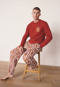 Rode shirt met lange mouwen en een afbeelding van een peperkoekman, gecombineerd met gestreepte pyjamabroek met herhaalde peperkoekmotieven in bruin en rood.