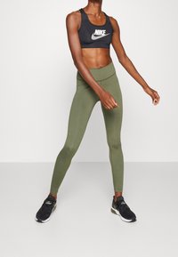 Sujeta deportiva negra con logo blanco de Nike, combinada con leggings de tiro alto en verde oliva. Modelo lleva zapatillas deportivas negras.