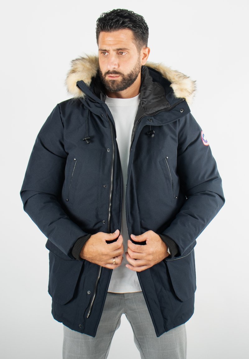 Parka marine avec capuche en fausse fourrure. Dotée d'une fermeture éclair, de poches latérales et de poignets élastiques. Fabriquée en tissu durable, semble isolée.