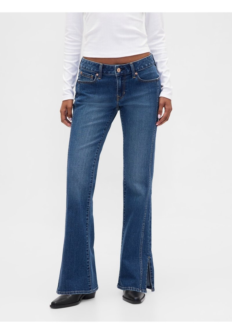 Femme portant un jean évasé bleu foncé avec des fentes latérales et des chaussures noires, assorti d'un haut court blanc à manches longues.