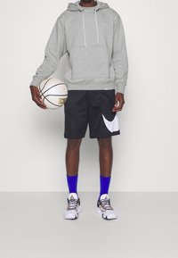 Homme portant un sweat à capuche gris, un short de basket noir, des chaussettes bleues et des baskets blanches, tenant un ballon de basket blanc sur un fond uni.