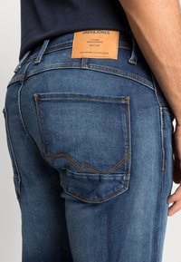 Siniset farkut oransseilla ompeleilla, joissa on nahkainen etiketti vyötäröllä, takataskuissa hienovaraisia geometrisia kuvioita ja slim fit -malli.