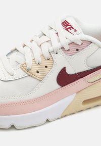 Nike Air Max tenisice s bijelom kožom, svijetloružičastim i bež naglascima, teksturiranim preklopima i bordo Swoosh logom.