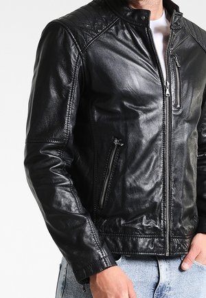 Mand iført en sort læderjakke med lynlåslommer over en hvid skjorte og lyseblå jeans, hånd hvilende nær lommen.