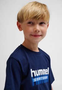 Mørkeblå bomuld t-shirt med et hvidt og blåt "hummel SPORTSWEAR" logo, korte ærmer og rund hals. Glat tekstur.