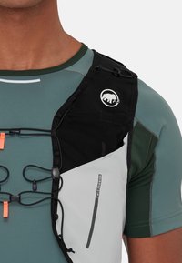 Black and grey running vest with adjustable straps, mesh pocket, elastic bungee cords, and logo. Worn over a green athletic shirt.