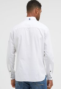 White long-sleeve shirt with a button-down collar, featuring black button accents on cuffs and a small blue label at the neckline.