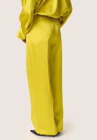 Pantaloni gialli a gamba larga in satin con una texture liscia, vita alta e pieghe sottili. Scarpe nere a punta completano l'outfit.