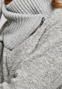 Grå stickad tröja med en ribbad turtleneck och en dragkedja som accent. Mjuk textur med varierade stickade mönster som visar upp en lagrad design.