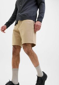 Shorts en coton beige avec un devant plat, des poches latérales et une coupe décontractée, associés à un cardigan sombre et des chaussures et chaussettes noires.