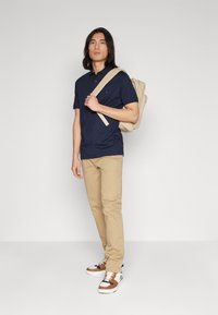 Marine polo shirt, beige broek en wit sneakers met bruine accenten. Model houdt een lichtbeige rugzak vast met verstelbare schouderbanden. Eenvoudige achtergrond.
