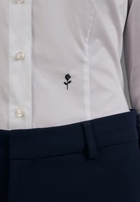 Chemise blanche boutonnée avec une petite broderie de rose noire près du bas devant, associée à un pantalon bleu marine foncé.