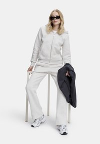 Lys grå bomull zip-up hettegenser og matchende joggebukser, kombinert med svarte solbriller. Hun holder en mørkegrå genser. Hvit bakgrunn.