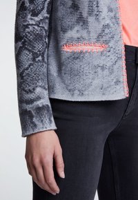 Cardigan gris tricoté avec un motif texturé et des détails de couture rose le long du bord ; porté sur un pantalon noir, affichant un style décontracté.