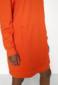 Personne portant une robe à manches longues orange vif avec des poches latérales, montrant une main et une partie des jambes nues sur un fond uni.