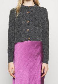 Šedý pletený cardigan s žebrovanou strukturou, zaobleným výstřihem a pěti dřevěnými knoflíky, nošený přes výrazné růžové saténové šaty.