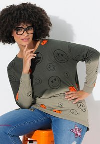 Blusa de manga larga verde y beige con patrones de caras sonrientes, combinada con jeans azules que tienen un parche de estrella con lentejuelas. Modelo sentada en un taburete naranja.