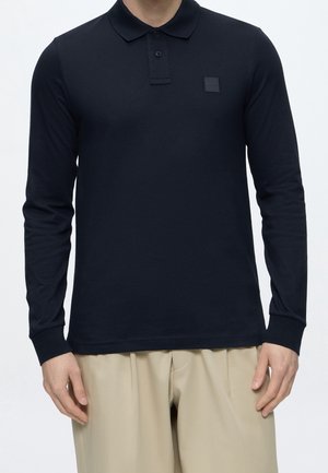 Mann trägt ein schwarzes langärmliges Poloshirt mit kleinem Logo auf der Brust und beigefarbenen Hosen vor reinem Hintergrund.