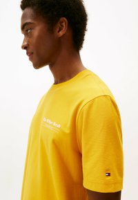 Junger Mann in einem gelben T-Shirt mit weißem Text auf der Brust und einem kleinen rot-weiß-blauen Logo am Ärmel, das nach links schaut vor einem einfarbigen Hintergrund.