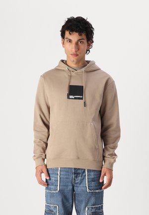 Beige Kapuzenpullover mit vorderer Kängurutasche, ausgestattet mit einem schwarzen Logo-Aufnäher. Aus weichem Stoff gefertigt, mit verstellbarem Kapuzenzug und gerippten Bündchen.