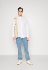 Camisa blanca de lino de botones con un corte relajado, combinada con jeans de color azul claro y zapatillas blancas. Lleva una bolsa cruzada acolchada beige.