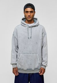 Lichtgrijze hoodie van zacht materiaal, met een voorzak, trekkoorden en gespatte zwarte details. Losse fit met geribbelde manchetten.