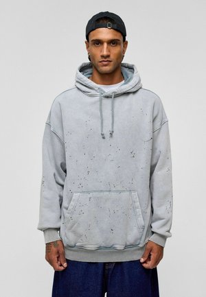 Lichtgrijze hoodie van zacht materiaal, met een voorzak, trekkoorden en gespatte zwarte details. Losse fit met geribbelde manchetten.