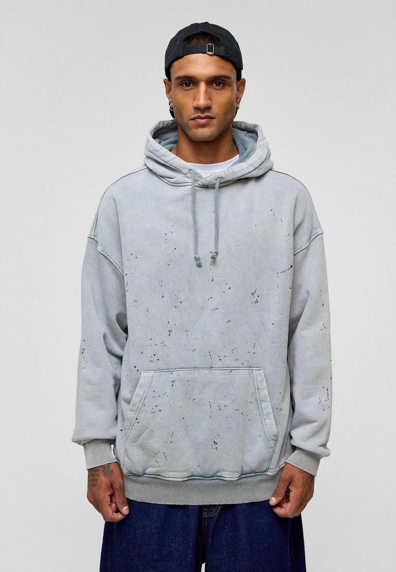 Lichtgrijze hoodie van zacht materiaal, met een voorzak, trekkoorden en gespatte zwarte details. Losse fit met geribbelde manchetten.