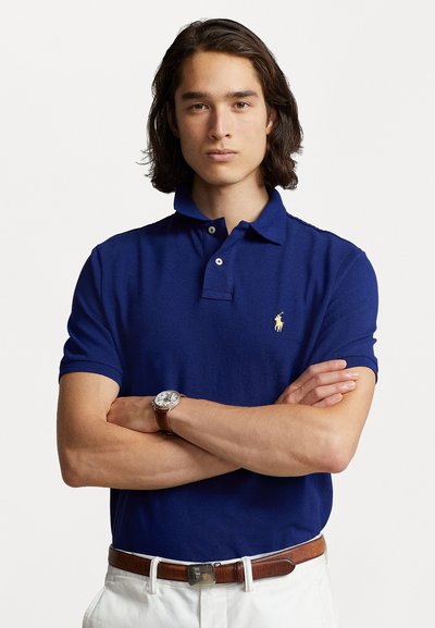 Polo bleu marine avec une finition texturée, patte de boutonnage à trois boutons et logo brodé jaune. Porté avec une montre en cuir marron et un pantalon blanc.