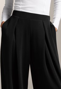 Fekete, bő szárú nadrág, redőkkel, sima anyagból készült. Oldalsó zsebekkel és lapos derékpánttal rendelkezik, fehér hosszú ujjú felsővel párosítva.