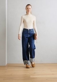 Beige, geripptes Langarmtop, hochgeschnittene, weite blaue Jeans mit hellem blauem Saum, braune Sneakers, hält eine kleine braune Handtasche.