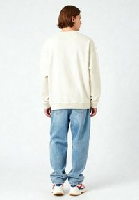 Felpa color crema con vestibilità rilassata, abbinata a jeans larghi azzurri. Il modello indossa sneakers bianche con dettagli rossi e blu.