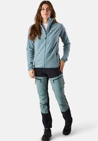 Vaaleansininen fleece-takki vetoketjulla, yhdistettynä kaksivärisiin housuihin vaaleansinisen ja laivastonsinisen väriyhdistelmässä, joissa on vetoketjulliset taskut ja vahvistetut polvet.