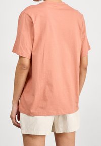 Luźna koszulka z okrągłym dekoltem, wykonana z miękkiej bawełny w kolorze koralowym. Krótkie rękawy, prosty dół i gładka faktura. Połączona z jasnobeżowymi szortami.