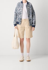 Bomber jacket con motivo floreale blu e bianco, t-shirt crema, pantaloni corti beige, sneakers bianche con suola marrone chiaro e una borsa a spalla crema.