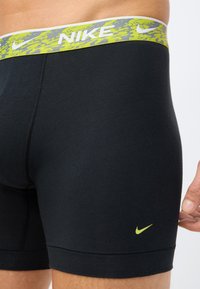 Čierne športové šortky s pásom v šedých a neonovo zelených vzoroch. Vyznačujú sa malým žltým logom Nike na boku a hladkou textúrou.