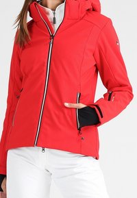 Rød vandtæt jakke med tekstureret mønster, lynlåslukning foran, justerbar hætte og sorte detaljer. Har to sidelommer og en tætsiddende pasform.
