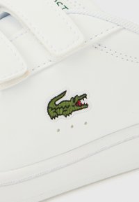 Λευκό δερμάτινο αθλητικό παπούτσι με λουριά Velcro, ραμμένες λεπτομέρειες, πράσινο κεντημένο λογότυπο κροκοδείλου και μικρές οπές για αερισμό.