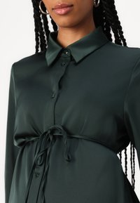 Σκούρο πράσινο, μεταξένιο μπλουζάκι με γιακά, κουμπί κλεισίματος και ζωνάκι στη μέση, με απαλό υφή και χαλαρή εφαρμογή.